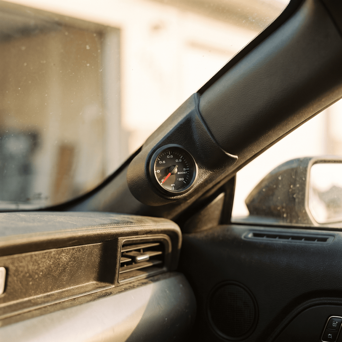 Black analog boost gauge mounted on car A-pillar with white face and red needle